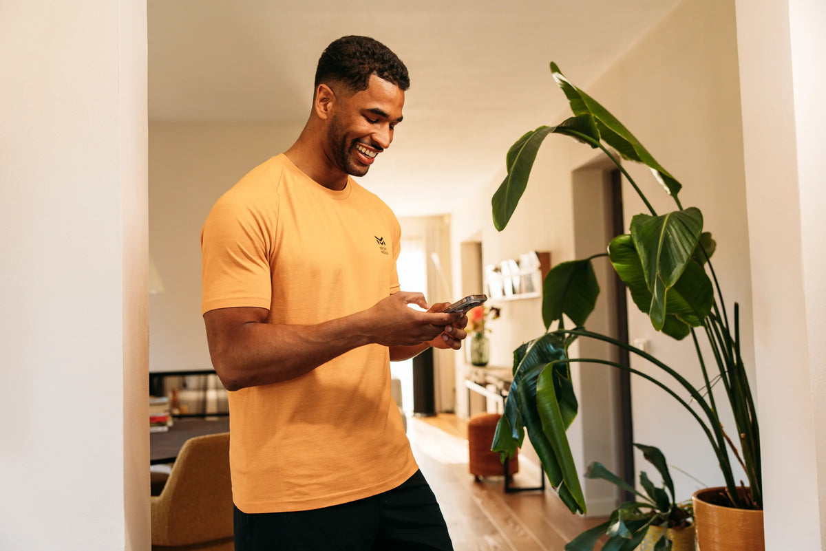 Sporter in een oranje SportMeals-shirt kijkt lachend op zijn telefoon in de woonkamer