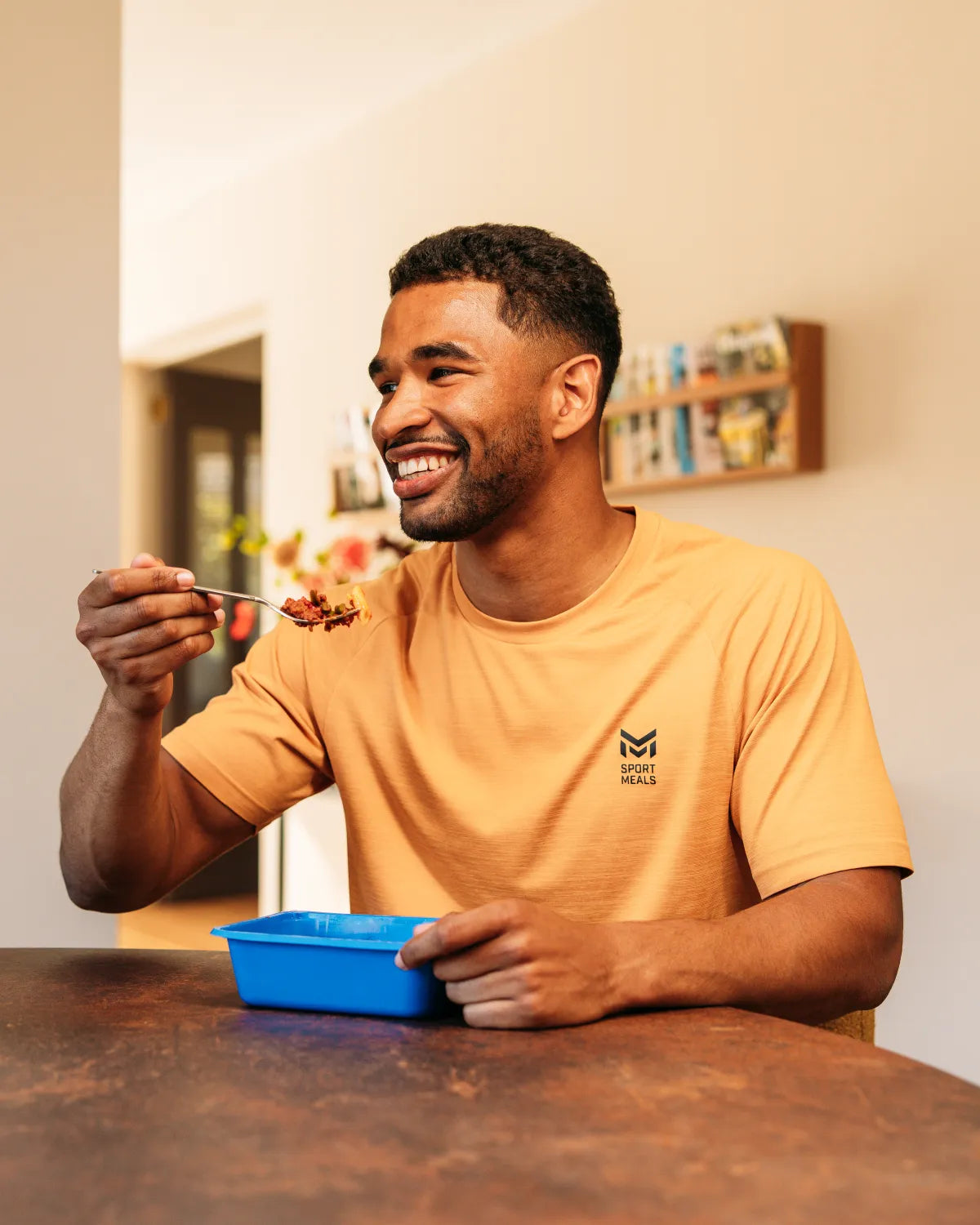Man in SportMeals-shirt eet lachend een maaltijd uit het blauwe bakje aan de eettafel