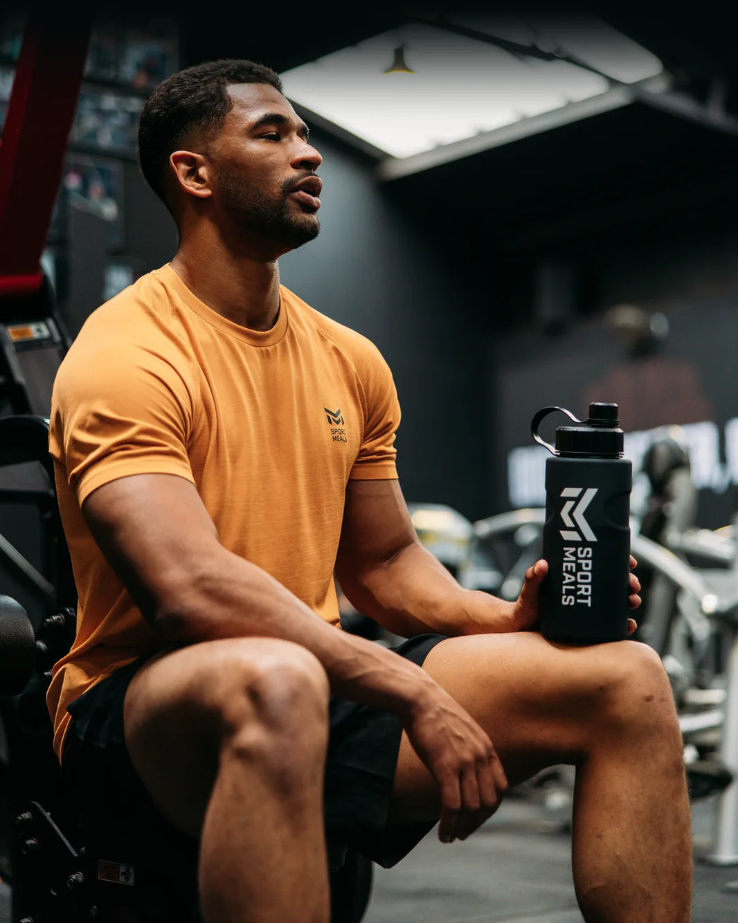 Bob in een oranje SportMeals-shirt zit in de sportschool met een SportMeals-drinkfles op zijn knie