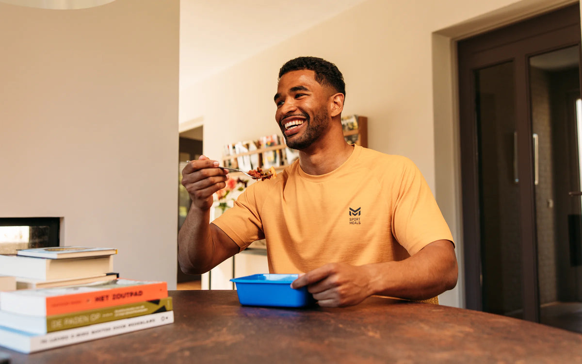 Een sporter eet lachend aan de eettafel uit een blauw SportMeals-bakje