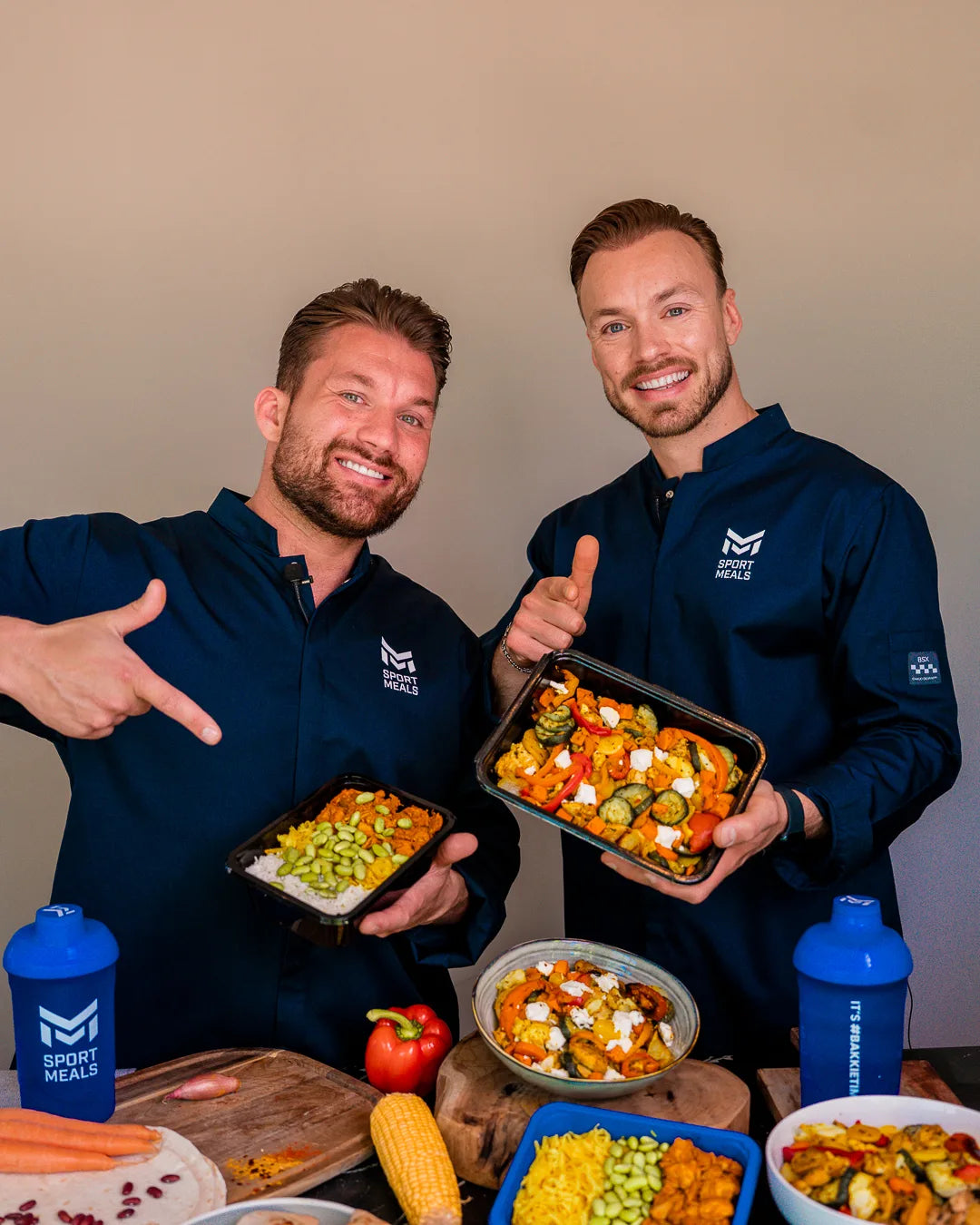 Joey en Ronald in koksbuis lachen in de camera; Joey wijst naar de kip Koreaans in zijn hand, Ronald houdt een traybake vast