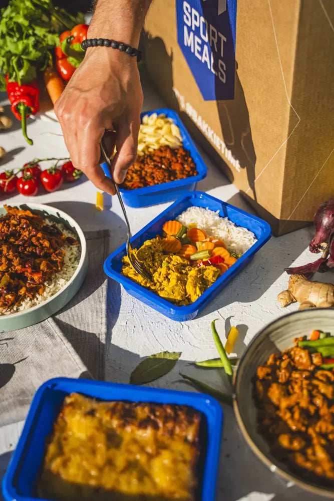 Een hand pakt met een vork een hap uit een blauwe SportMeals-maaltijd op tafel