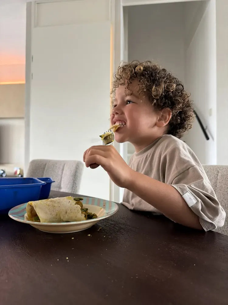 A young child smiles while taking a bite at the table, with a blue SportMeals meal in the background