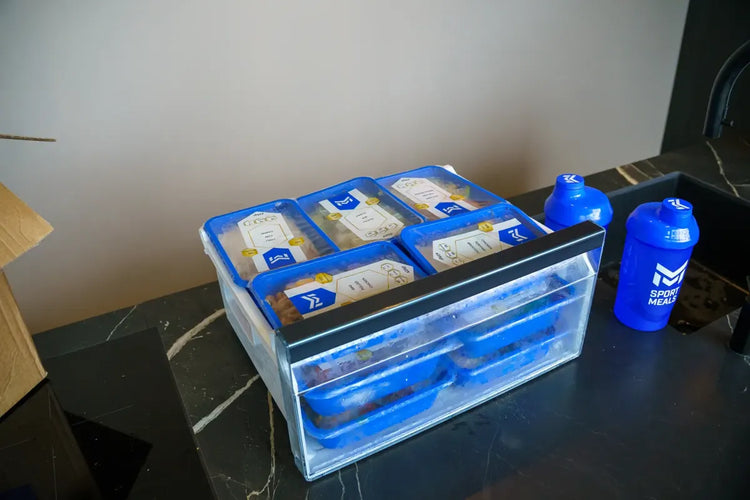 Een vriezerlade vol gestapelde SportMeals-maaltijden naast een blauwe shaker
