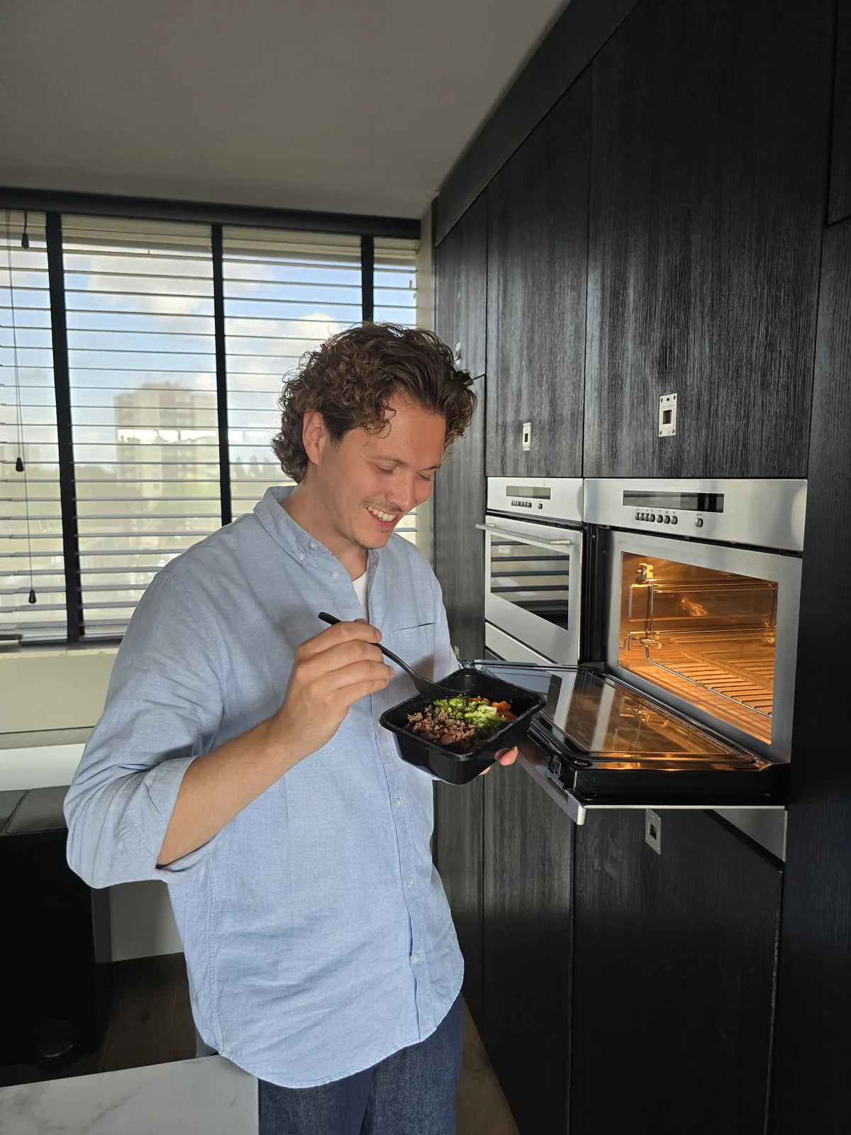 Delano stands smiling in the kitchen next to the open oven, eating from a SportMeals meal with a fork