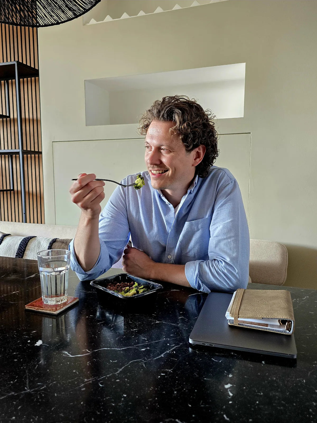 Delano sits at the table smiling and takes a bite of a SportMeals meal, with his laptop closed next to him