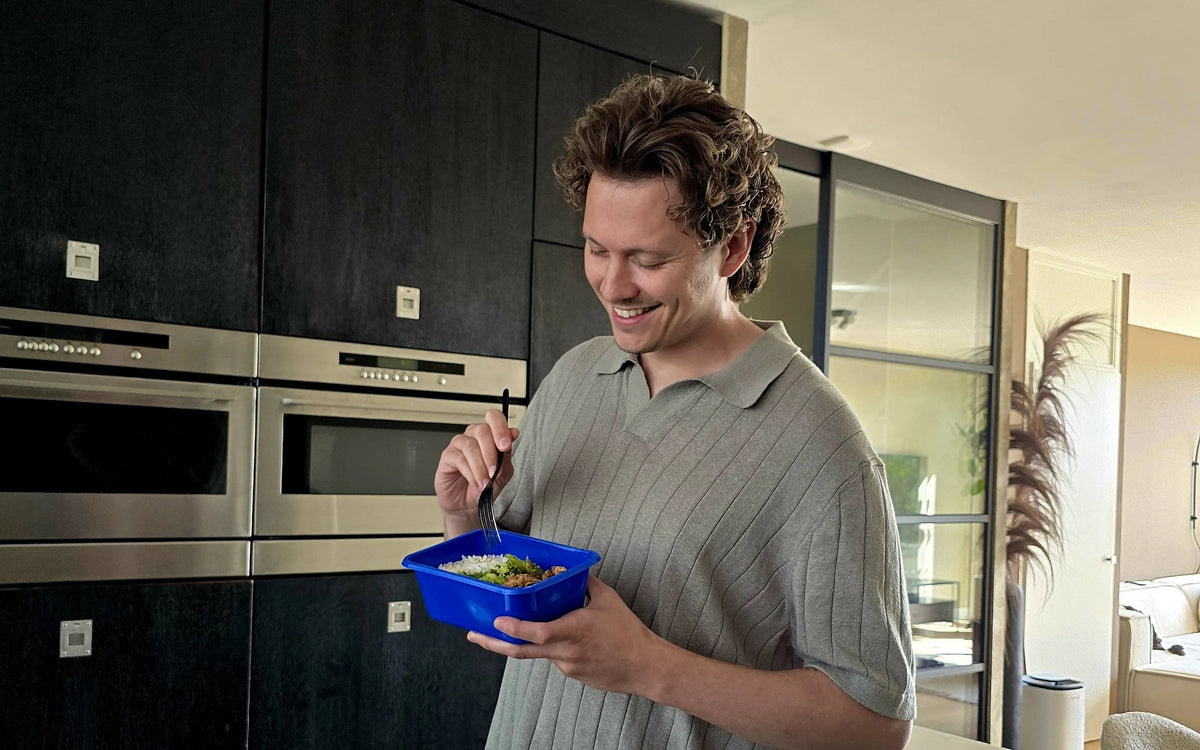Delano stands smiling in the kitchen, eating with a fork from a blue SportMeals meal