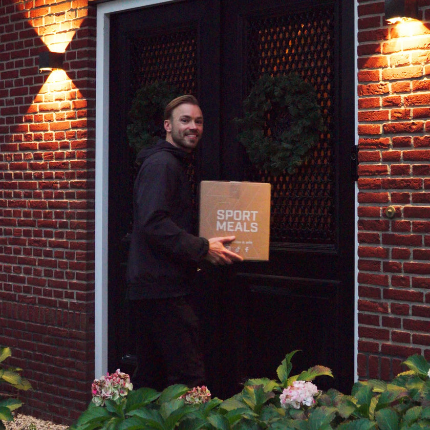 Man staat ’s avonds lachend bij de voordeur met een SportMeals-doos in zijn handen