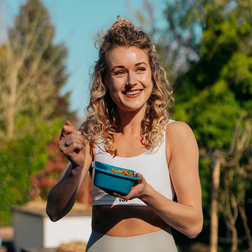 Vrouw eet buiten lachend uit een blauw SportMeals-bakje